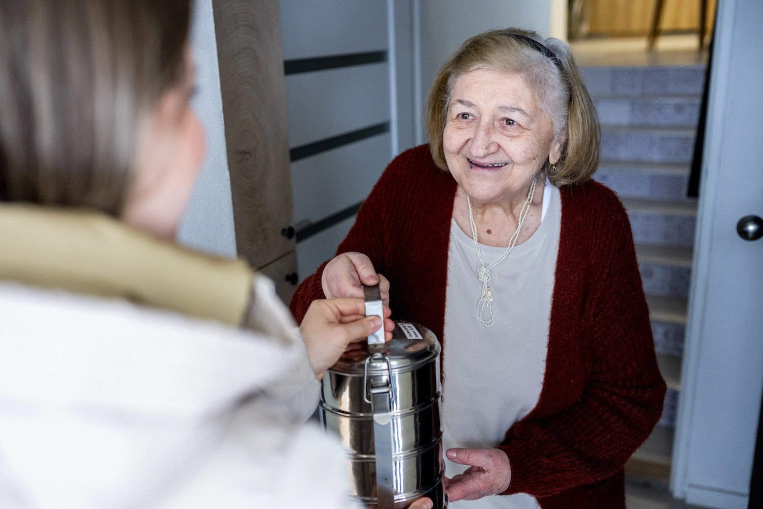 A GA Foods driver hands a stainless steel meal container to a smiling senior woman at her front door — meal delivery route optimization in action
